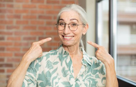 Téléchargez les photos : Senior jolie femme souriant avec confiance pointant vers son propre sourire large, attitude positive, détendue, satisfaite - en image libre de droit