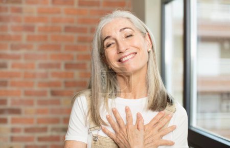 Téléchargez les photos : Senior jolie femme se sentant romantique, heureux et amoureux, souriant joyeusement et tenant les mains près du cœur - en image libre de droit
