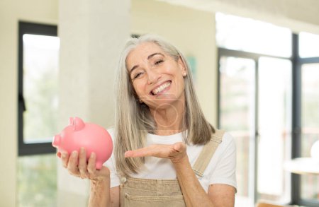 Photo for Pretty senior woman smiling cheerfully, feeling happy and showing a concept. with a piggy bank - Royalty Free Image