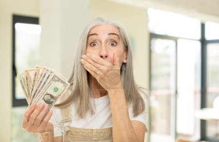 Photo for Pretty senior woman covering mouth with a hand and shocked or surprised expression. with dollar banknotes - Royalty Free Image