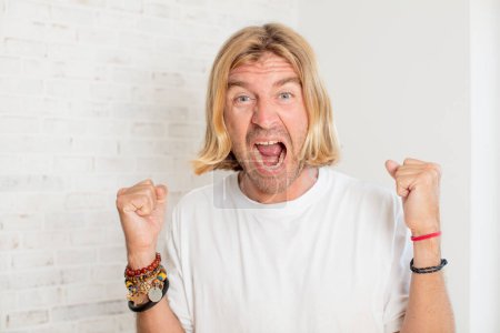 Photo for Young blond adult man feeling shocked, excited and happy, laughing and celebrating success, saying wow! - Royalty Free Image