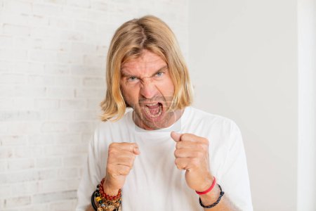 Photo for Young blond adult man shouting aggressively with annoyed, frustrated, angry look and tight fists, feeling furious - Royalty Free Image