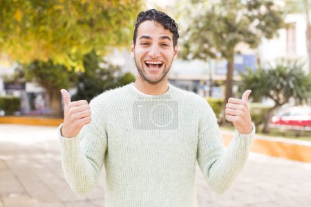 Foto de Joven hispano sonriendo alegremente y luciendo feliz, sintiéndose despreocupado y positivo con ambos pulgares arriba - Imagen libre de derechos