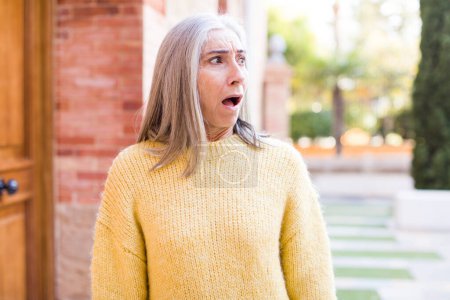 Photo for Senior retired pretty white hair woman feeling shocked and surprised, looking to copy space on the side with amazed, open-mouthed look - Royalty Free Image