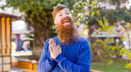 Foto de Hombre barbudo de pelo rojo sintiéndose feliz y exitoso, sonriendo y aplaudiendo, diciendo felicitaciones con un aplauso - Imagen libre de derechos
