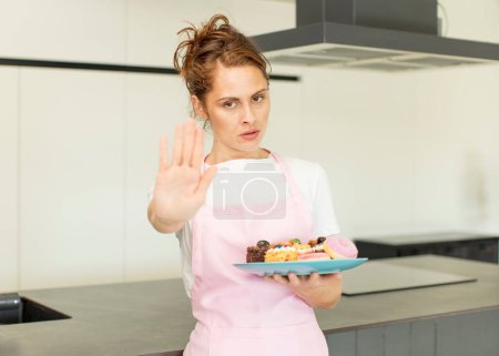 Téléchargez les photos : Young pretty woman looking serious showing open palm making stop gesture. home made cakes concept - en image libre de droit