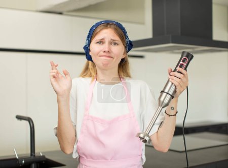Foto de Joven mujer bonita cruzando los dedos y esperando buena suerte. concepto de chef y licuadora de mano - Imagen libre de derechos