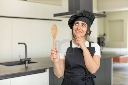 Foto de Mujer joven sonriendo con una expresión feliz y segura con la mano en la barbilla. concepto de chef - Imagen libre de derechos