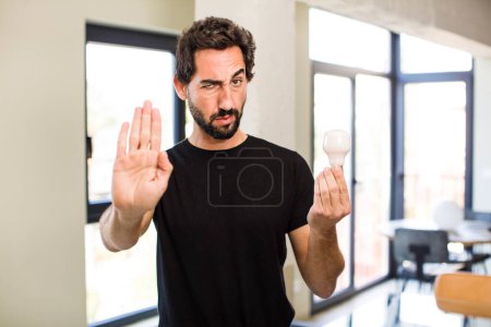 Foto de Joven barbudo loco mirando serio mostrando palmera abierta haciendo gesto de parada con una bombilla - Imagen libre de derechos
