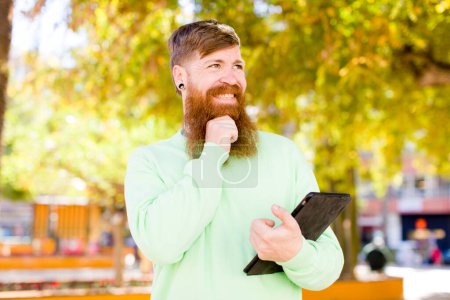 Téléchargez les photos : Cheveux roux jeune homme adulte souriant avec une expression heureuse et confiante avec la main sur le menton avec un tampon tactile - en image libre de droit