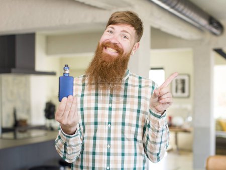 Photo for Red hair man smiling cheerfully, feeling happy and pointing to the side with a vaper - Royalty Free Image