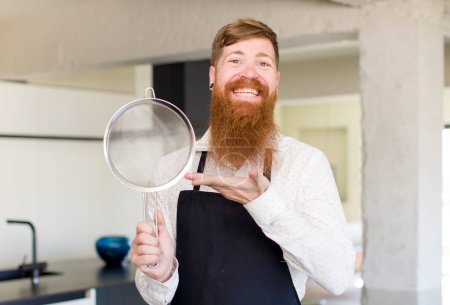 Foto de Hombre de pelo rojo sonriendo alegremente, sintiéndose feliz y mostrando un concepto en una cocina. concepto de chef - Imagen libre de derechos