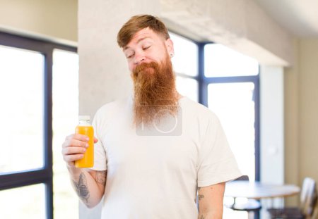 Foto de Hombre de pelo rojo sonriendo y mirando con una expresión feliz y segura con una botella de jugo de naranja - Imagen libre de derechos