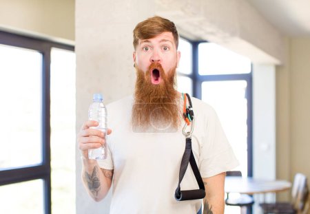 Foto de Hombre de pelo rojo se siente muy sorprendido y sorprendido con una botella de agua. concepto de fitness - Imagen libre de derechos