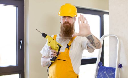 Téléchargez les photos : Homme aux cheveux roux regardant sérieusement montrant la paume ouverte faisant geste d'arrêt réparer la maison. concept bricoleur - en image libre de droit