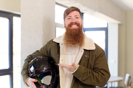 Téléchargez les photos : Homme aux cheveux roux souriant joyeusement, se sentant heureux et montrant un concept avec un casque de moto - en image libre de droit