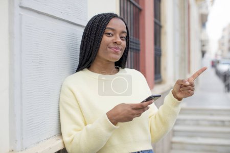Foto de Negro afro mujer sonriendo alegremente, sintiéndose feliz y señalando hacia un lado. usando un concepto de smartphone - Imagen libre de derechos