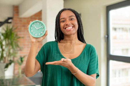 Photo for Black afro woman smiling cheerfully, feeling happy and showing a concept. alarm clock concept - Royalty Free Image