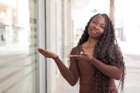 Foto de Mujer negra bonita afro sonriendo con orgullo y confianza, sintiéndose feliz y satisfecho y mostrando un concepto en el espacio de copia - Imagen libre de derechos
