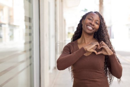Téléchargez les photos : Afro jolie femme noire souriante et se sentant heureuse, mignonne, romantique et amoureuse, faisant forme de coeur avec les deux mains - en image libre de droit