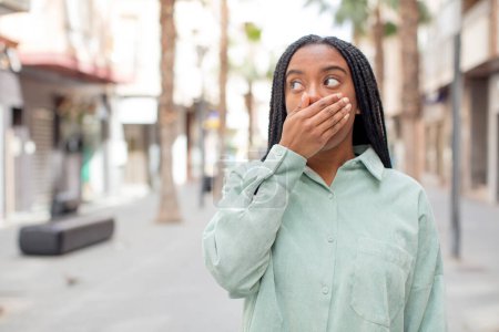 Téléchargez les photos : Afro jolie femme noire couvrant la bouche avec les mains d'une expression choquée et surprise, gardant un secret ou disant oups - en image libre de droit