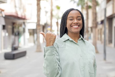 Téléchargez les photos : Afro jolie femme noire souriant joyeusement et décontracté pointant vers copier l'espace sur le côté, se sentant heureux et satisfait - en image libre de droit