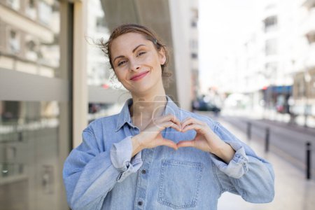 Photo for Pretty young woman smiling and feeling happy, cute, romantic and in love, making heart shape with both hands - Royalty Free Image