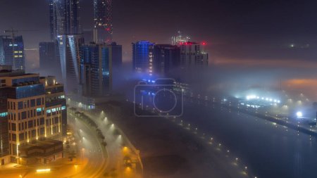 Foto de Los edificios están cubiertos de una gruesa capa de niebla en el lapso de tiempo nocturno de Business Bay. Rascacielos iluminados alrededor del canal de agua vista panorámica aérea - Imagen libre de derechos