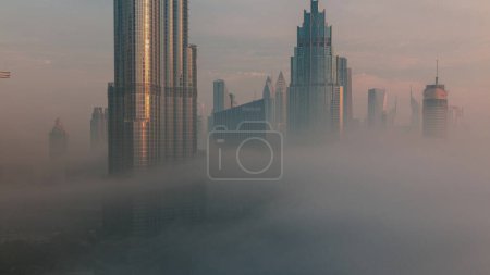 Foto de Vista aérea de la ciudad de Dubai temprano en la mañana durante el timelapse niebla. Salida del sol en el horizonte futurista de la ciudad con rascacielos y torres desde arriba. Sol reflejado desde la superficie de vidrio - Imagen libre de derechos
