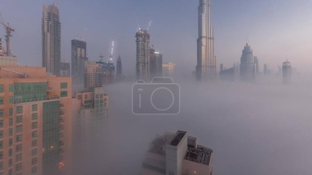 Téléchargez les photos : Vue aérienne de la ville de Dubaï tôt le matin pendant la nuit de brouillard à l'heure du jour. Skyline futuriste de la ville avec des gratte-ciel et des tours en construction d'en haut - en image libre de droit