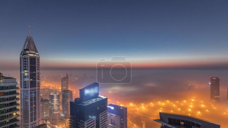 Téléchargez les photos : Brouillard hivernal rare tôt le matin au-dessus de la ligne d'horizon de Dubai Marina et gratte-ciel éclairés par des lampadaires aériens nuit à jour passage timelapse. Vue de dessus depuis les nuages. Dubai, Émirats arabes unis - en image libre de droit