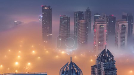 Photo for JLT skyscrapers near Sheikh Zayed Road aerial night to day transition timelapse. Illuminated residential buildings and traffic on crossroad junction. Foggy morning before sunrise - Royalty Free Image