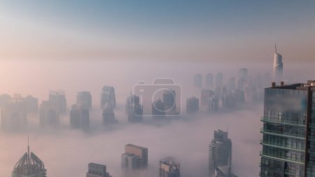 Téléchargez les photos : Le brouillard couvrait les gratte-ciel et les tours de marina de la JLT près de Sheikh Zayed Road après le lever du soleil. Immeuble résidentiel matin brumeux avec lumière orange chaude - en image libre de droit