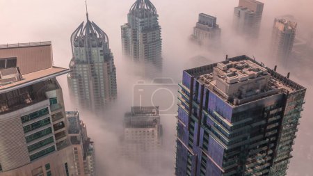 Téléchargez les photos : Tours couvertes de brouillard hivernal rare tôt le matin au-dessus de la ligne d'horizon de Dubai Marina et gratte-ciel sur les toits timelapse aérien. Vue de dessus depuis les nuages. Dubai, Émirats arabes unis - en image libre de droit