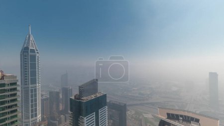 Téléchargez les photos : Tours couvertes de brouillard hivernal rare tôt le matin au-dessus de l'horizon de Dubai Marina avec soleil se levant et gratte-ciel sur les toits timelapse aérienne. Vue de dessus depuis les nuages. Dubai, Émirats arabes unis - en image libre de droit