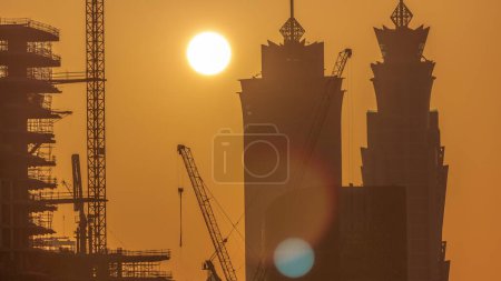 Téléchargez les photos : Vue rapprochée sur les tours de la baie d'affaires de Dubaï au coucher du soleil. Vue sur le toit de certains gratte-ciel et de nouvelles tours en construction. Ciel orange avec bâtiments silhouette et grues - en image libre de droit