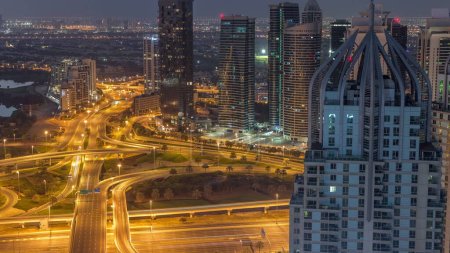 Téléchargez les photos : Un énorme carrefour routier entre le quartier JLT et la marina de Dubaï croisé par Sheikh Zayed Road, la nuit aérienne et le temps de transition diurne. Terrain de golf près des tours éclairées et des gratte-ciel avec un trafic achalandé - en image libre de droit