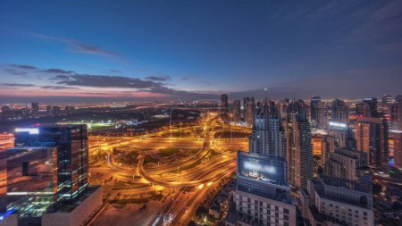 Téléchargez les photos : Énorme carrefour routier entre le quartier JLT et Dubai Marina intersecté par Sheikh Zayed Road transition aérienne nuit à jour timelapse panoramique. Terrain de golf près des tours éclairées et des gratte-ciel avec un trafic achalandé - en image libre de droit
