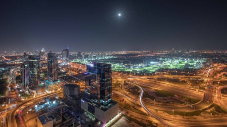Foto de Panorama que muestra la ciudad de los medios de comunicación, el puerto deportivo de Dubái y los rascacielos iluminados JLT a lo largo de Sheikh Zayed Road con gran cruce de cruce aéreo nocturno timelapse. Luna naciente sobre edificios residenciales y de oficinas y campo de golf desde arriba. - Imagen libre de derechos
