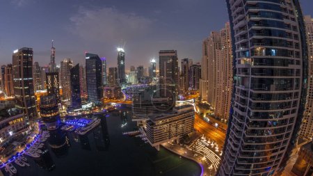 Téléchargez les photos : Vue aérienne du port de plaisance de Dubaï illuminait les gratte-ciel autour du canal avec des yachts flottants nuit à jour transition timelapse panorama. Les bateaux blancs sont garés dans le yacht club avant le lever du soleil - en image libre de droit
