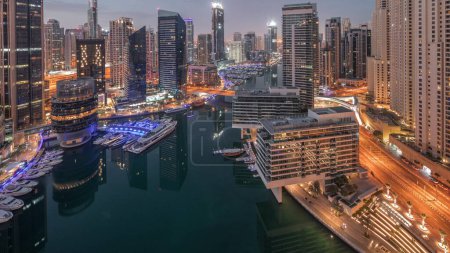 Téléchargez les photos : Vue aérienne sur la marina de Dubaï et les gratte-ciel jbr illuminés autour du canal avec des yachts flottants nuit à jour passage timelapse. Les bateaux blancs sont garés dans le yacht club avant le lever du soleil - en image libre de droit