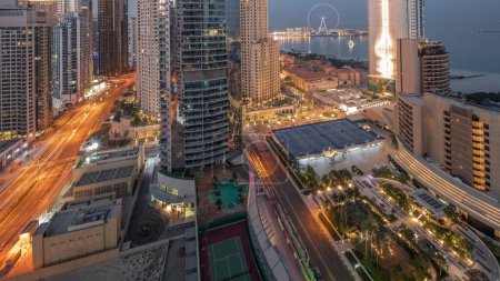Téléchargez les photos : Vue panoramique sur le port de plaisance de Dubaï et la région de JBR et la célèbre roue Ferris nuit aérienne à la transition de jour timelapse. gratte-ciel lumineux au lever du soleil avec de longues ombres - en image libre de droit