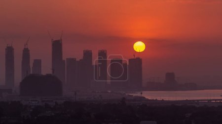 Foto de Salida del sol sobre Dubai Creek Harbor con rascacielos y torres en construcción timelapse aéreo. Gran Sol rojo se levanta detrás de los edificios - Imagen libre de derechos