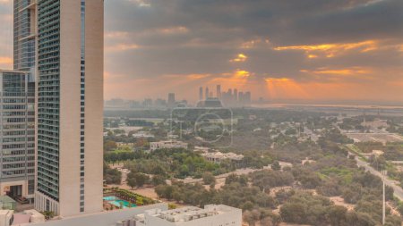 Foto de Salida del sol sobre el jardín en el distrito de Zabeel con rascacielos en un timelapse aéreo de fondo en Dubai, Emiratos Árabes Unidos. Nubes anaranjadas en el cielo con rayos de luz solar - Imagen libre de derechos