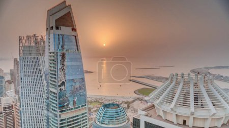 Téléchargez les photos : Vue aérienne du coucher du soleil sur les gratte-ciel et les bâtiments de luxe de JBR et Dubai Marina timelapse d'en haut. Tours modernes avec plage et soleil sur ciel orange reflété dans la mer - en image libre de droit