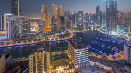 Foto de Dubai Marina con barcos y yates estacionados en el puerto y rascacielos iluminados alrededor del canal aéreo noche a día timelapse transición antes del amanecer. Torres del distrito JBR en segundo plano - Imagen libre de derechos