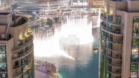 Téléchargez les photos : Fontaines chantantes de Dubaï avec zone de marche autour de la vue aérienne de nuit entre les gratte-ciel timelapse. Architecture avec maisons traditionnelles dans la vieille ville. Les gens regardent le spectacle près du centre commercial - en image libre de droit
