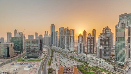 Téléchargez les photos : Bay Avenue au coucher du soleil avec des tours modernes développement résidentiel à Business Bay ciel panoramique timelapse, Dubaï, Émirats arabes unis. gratte-ciel avec circulation sur une route près d'un grand parking - en image libre de droit