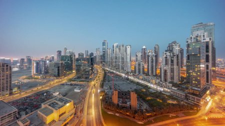 Foto de Bay Avenue con torres modernas desarrollo residencial en Business Bay noche aérea al día panorama timelapse transición, Dubai, Emiratos Árabes Unidos. Rascacielos con tráfico en una carretera cerca de un gran aparcamiento - Imagen libre de derechos