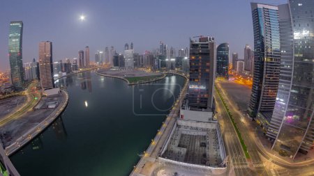 Téléchargez les photos : Paysage urbain panorama de gratte-ciel dans la baie d'affaires de Dubaï avec canal d'eau de nuit aérienne au jour de transition timelapse avec la lune couchant. Skyline moderne avec tours éclairées et front de mer avant le lever du soleil - en image libre de droit